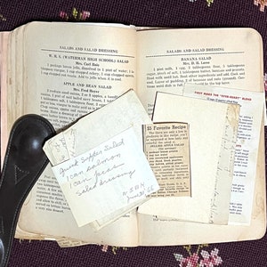 May include: An open vintage cookbook with handwritten recipe cards and printed salad recipes. The pages are yellowed with age, and a black leather spoon rests on the side. The recipe cards include "Quick Supper Salad" and "Jellied Apple Salad".
