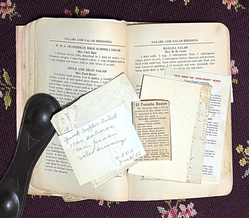 May include: An open vintage cookbook with handwritten recipe cards and printed salad recipes. The pages are yellowed with age, and a black leather spoon rests on the side. The recipe cards include "Quick Supper Salad" and "Jellied Apple Salad".