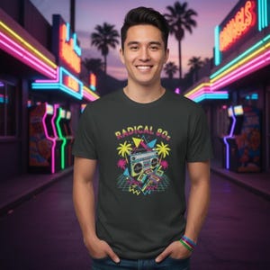 Puede incluir: Camiseta gris oscuro con un gráfico colorido de los años 80. El diseño presenta una radio, palmeras y el texto "RADICAL 80s" en una fuente retro. La persona lleva la camiseta frente a una sala de juegos de neón.