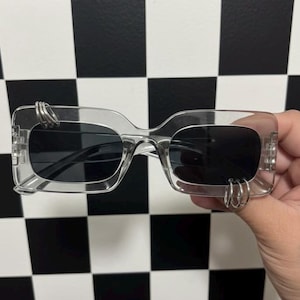 May include: Clear rectangular sunglasses with dark lenses and silver ring accents on the frames. The glasses have a transparent frame and are set against a black and white checkered background.