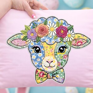 May include: A pink cushion with a colourful lamb design. The lamb's face is made of patchwork fabric with floral patterns, a floral crown, and a bow tie. The design is outlined with a beaded trim.