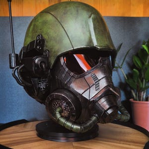 Pode incluir: Um capacete tático com uma cúpula verde-oliva desgastada e uma placa frontal metálica escura com uma viseira avermelhada. O capacete inclui uma antena montada na lateral, componentes mecânicos pretos e um respirador frontal com duas mangueiras.