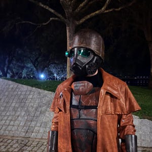 Op de afbeelding: Een bruine en zwarte post-apocalyptische helm en masker met groene lenzen. De persoon draagt een bruine leren jas en een borstplaat met de tekst "GT VICKERS OPOS USMC". Het kostuum staat tegen een donkere achtergrond.