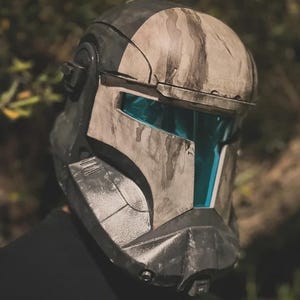 May include: A detailed, weathered helmet with a blue visor. The helmet is primarily gray and white with dark gray accents and a camouflage pattern. The design suggests a futuristic or science fiction theme, possibly for cosplay or display.