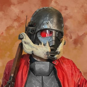 Puede incluir: Un casco táctico gris oscuro con una visera roja y una pieza de mandíbula ósea que sobresale en la parte frontal. El casco cuenta con accesorios laterales y se lleva con una chaqueta roja brillante sobre una placa de pecho gris oscuro y un rifle.