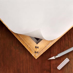 May include: A light-colored wooden picture frame corner with a white paper sheet partially peeled back to reveal a dark interior. A gray craft knife with a removable cap is positioned nearby on the dark wood surface.