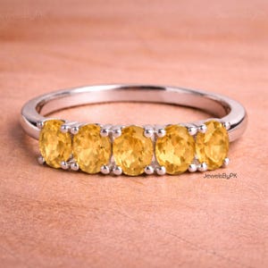 Op de afbeelding: Een zilveren ring met vijf ovale, goudgele edelstenen. De edelstenen zijn naast elkaar geplaatst. De ring heeft een gladde, gepolijste band. De achtergrond is lichtbruin.