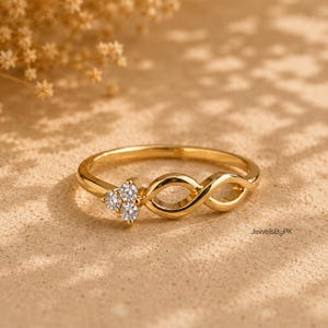 Pode incluir: Um anel de ouro com uma banda polida, apresentando um design de símbolo de infinito num dos lados e um grupo de três pequenas pedras redondas, claras e facetadas, dispostas em triângulo na extremidade do laço do infinito.