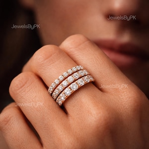 May include: Three rose gold eternity rings, each encrusted with numerous small, sparkling diamonds. The rings are stacked on a finger, showcasing their design. The image has the text "JewelsByPK" in the corners.