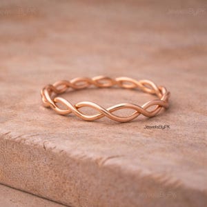 Può includere: Anello in oro rosa con un design a torsione infinita. L'anello è realizzato in metallo lucido e poggia su una superficie marrone chiaro. Il design dell'anello presenta un motivo a ciclo continuo, creando un aspetto delicato ed elegante. È un gioiello.