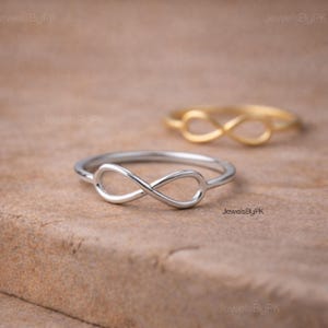 Pode incluir: Dois anéis de infinito, um prateado e outro dourado, são exibidos em uma superfície texturizada. Os anéis, com o símbolo do infinito, são o foco principal. O anel prateado está em foco nítido, enquanto o anel dourado está ligeiramente desfocado no fundo.