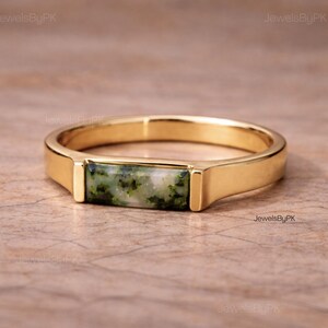 May include: A gold-colored ring with a rectangular, moss agate stone. The stone features green and white patterns. The ring has a simple, modern design, with a polished finish. The ring is a piece of jewelry.