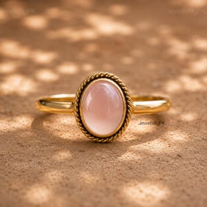 Pode incluir: Um anel de ouro com uma pedra preciosa lisa, de cor rosa pálido e formato oval no centro. A pedra é rodeada por uma borda metálica decorativa com um design de corda torcida. O aro do anel é fino, polido e de aparência dourada.