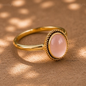 Op de afbeelding: Een gouden ring met een gladde, ovaalvormige, lichtroze cabochon edelsteen in een zetting. De zetting is omringd door een decoratieve gedraaide metalen rand in touwstijl, en de ringband heeft een eenvoudige, gepolijste gouden afwerking.