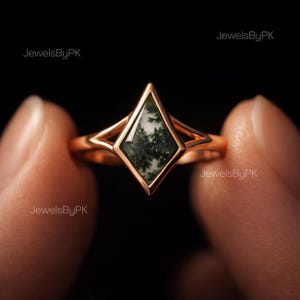 May include: A rose gold ring with a diamond-shaped moss agate stone. The stone displays a green and white pattern, set in a delicate, open-work frame. The ring is held against a dark background.