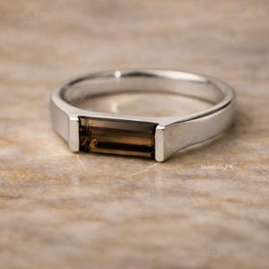 Könnte beinhalten: Ein silberner Ring mit einem rechteckigen, braunen Edelstein. Der Ring hat ein schlichtes, modernes Design mit poliertem Finish. Der Edelstein ist in einer Lünettenfassung gefasst, und das Band ist glatt und abgerundet.