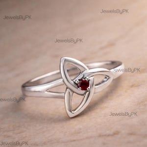 May include: A silver ring with a Celtic knot design, featuring a central, round, deep red gemstone. The ring has a smooth, polished band, and the knotwork is open. The ring is set against a light brown background.