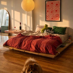 Könnte beinhalten: Ein niedriges Holzplattformbett verfügt über eine tiefrote, strukturierte Bettdecke, die mit beigen und grünen Kissen geschichtet ist. Ein flauschiger brauner Hund sitzt auf dem Hartholzboden vor dem Bett. Eine runde Papierpendelleuchte hängt darüber und ein rot-weißer abstrakter Druck ist an der Wand befestigt.