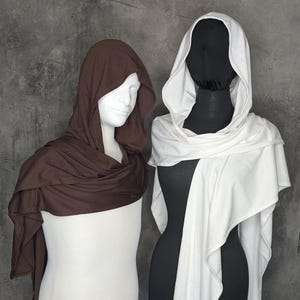 May include: Two hooded cowls displayed on mannequins. One cowl is solid dark brown, and the other is solid white. Both garments feature a draped, wrap-around design with an attached hood, constructed from a smooth, lightweight fabric material.