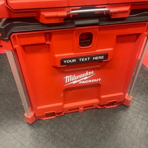 May include: Red Milwaukee Packout storage container with the text "YOUR TEXT HERE" and the Milwaukee logo. The container has a black handle and metal support bars. The container is sitting on a black surface.