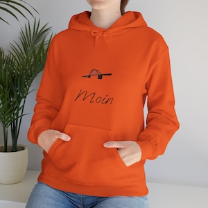 Puede incluir: Sudadera con capucha de color naranja brillante con bolsillo delantero y capucha con cordón. La sudadera presenta un gráfico negro de un puente y la palabra "Moin" en una fuente manuscrita. La persona lleva vaqueros azules.