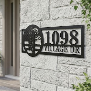 May include: Black metal address sign with the numbers "1098" and the words "VILLAGE DR". The sign has a decorative element featuring a design with a utility pole and American flag. It is mounted on a light-colored stone wall.