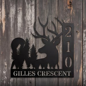 May include: A black metal house sign featuring a silhouette of a deer head, a forest of pine trees, and a circular moon. The right side displays the vertical numbers 210, and the bottom section features the text GILLES CRESCENT in capital letters.