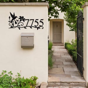 Puede incluir: Un letrero de metal negro con el número de casa 7755, que presenta una silueta decorativa de un colibrí en vuelo y detalles de enredaderas florales. El letrero está montado en una pared de color claro sobre un buzón de metal.