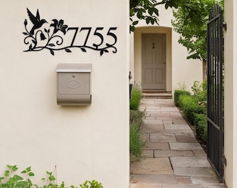 May include: A black metal house number sign displaying the digits 7755, featuring a decorative silhouette of a hummingbird in flight and floral vine accents. The sign is mounted on a light-colored wall above a metal mailbox.