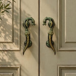 May include: A pair of vertical door handles featuring a curved, elongated design with intricate embossed detailing. The metal hardware displays a dark, weathered finish with highlighted gold and green accents, mounted on a light-colored door.