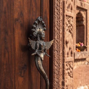 May include: A dark bronze-toned metal door handle shaped like a winged figure with a serpent-like tail and a multi-headed cobra hood, mounted vertically on a dark brown wooden door with a visible wood grain pattern and a carved stone frame.