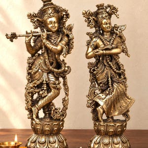 Puede incluir: Dos estatuas ornamentadas de color latón que representan deidades sobre pedestales de loto. La figura de la izquierda sostiene una flauta, mientras que la de la derecha tiene las manos en un gesto elegante. Ambas lucen joyas detalladas y coronas.