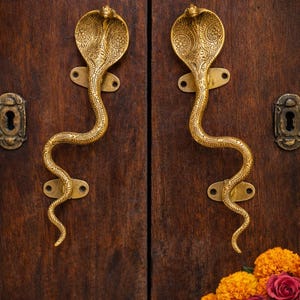 May include: Two brass cobra-shaped door handles mounted on a dark wooden door. The handles are detailed with engraved patterns and have a golden finish. The door also features keyholes and decorative plates.