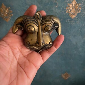 Op de afbeelding: Een messingkleurige, gestileerde sculptuur van een gezicht met opvallende kenmerken, waaronder grote ogen, een lange neus en een gedetailleerd voorhoofd. De sculptuur heeft een getextureerd oppervlak en wordt in een hand gehouden tegen een turquoise achtergrond.