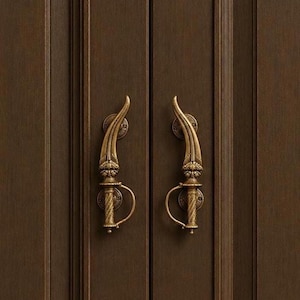 May include: Two antique brass door handles shaped like curved daggers with intricate engravings and a circular guard. The handles are mounted vertically on a dark brown wooden door with vertical paneling and a smooth, matte finish.