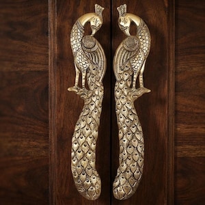 May include: A pair of brass door handles shaped like peacocks with intricate engraved feather details. The metallic gold-toned handles feature a bird standing on a branch with a long, textured tail extending downward in a vertical orientation.