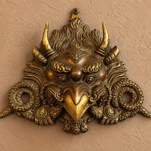 May include: A metallic gold-toned wall plaque featuring a detailed relief of a mythical creature with a bird-like beak, prominent horns, and swirling patterns. The design includes two circular openings on the sides and a single screw hole at the top.