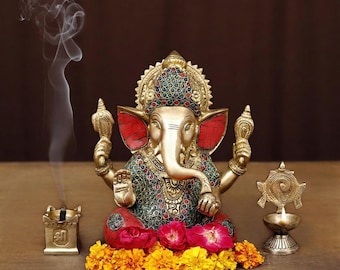 May include: A gold-colored statue of the Hindu deity Ganesha, adorned with colorful jewels and seated on a bed of flowers. Incense smoke rises from a small holder. A small lamp is lit nearby.