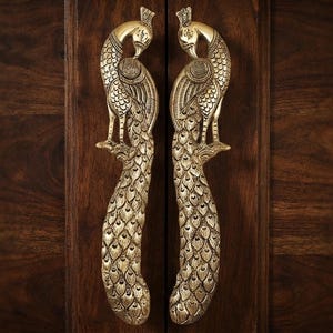 May include: A pair of vertical door handles crafted from polished brass, each featuring a detailed peacock design. The birds are depicted standing with their long, textured tail feathers extending downwards, showcasing intricate engraved patterns.
