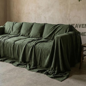 May include: An olive green sofa draped with a matching textured throw. The room features a neutral-toned wall and a window with sheer curtains. A wooden chair is positioned to the right of the sofa.