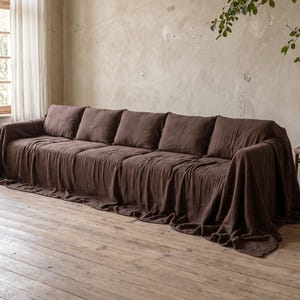 Może przedstawiać: Długa sofa przykryta narzutą z ciemnobrązowej, fakturowanej tkaniny, która zwisa przez podłokietniki aż do podłogi. Sofa posiada cztery duże, kwadratowe poduszki oparciowe oraz ciągłą poduszkę siedziska, wszystko w głębokim brązowym kolorze.