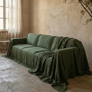 May include: A green sofa covered with a textured throw. The room has neutral walls and a window with sheer curtains. A wooden chair is positioned near the window. The sofa is approximately 200 cm in length.