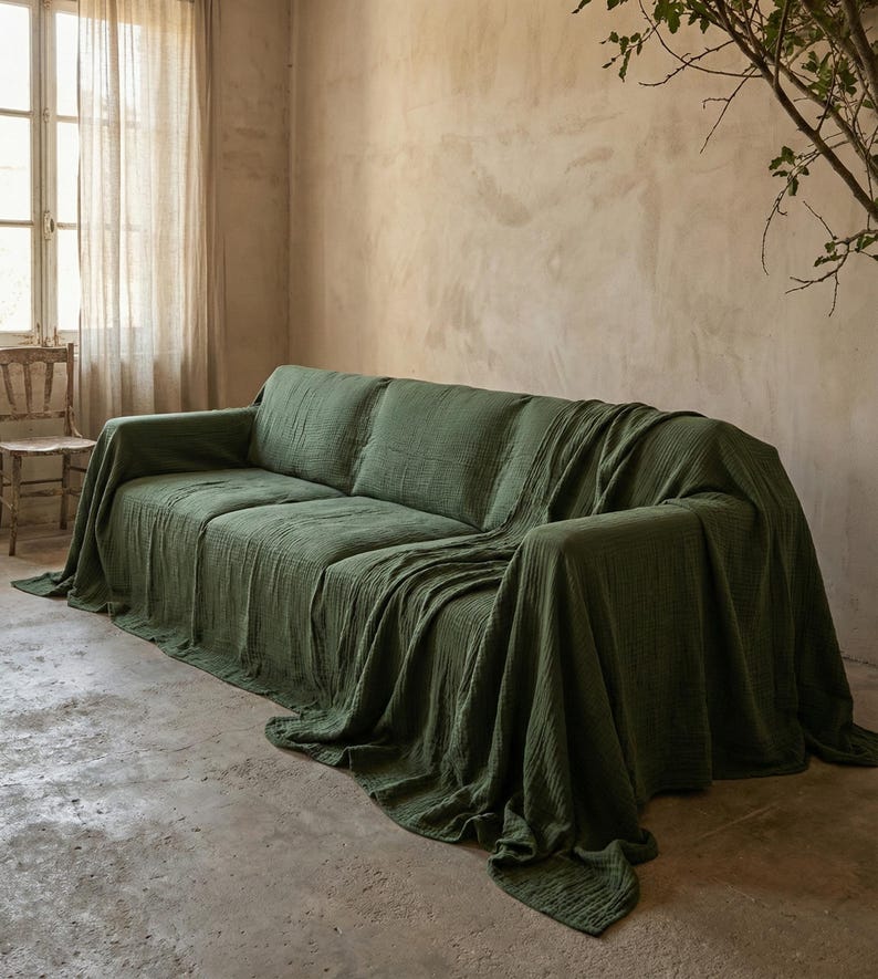 May include: A green sofa covered with a textured throw. The room has neutral walls and a window with sheer curtains. A wooden chair is positioned near the window. The sofa is approximately 200 cm in length.