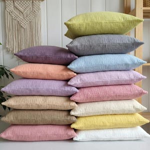 May include: A stack of fifteen rectangular throw pillows in various pastel colors, including yellow, gray, purple, peach, blue, and pink. The pillows have a textured, crinkled appearance, suggesting a soft fabric. The pillows are stacked on top of each other.