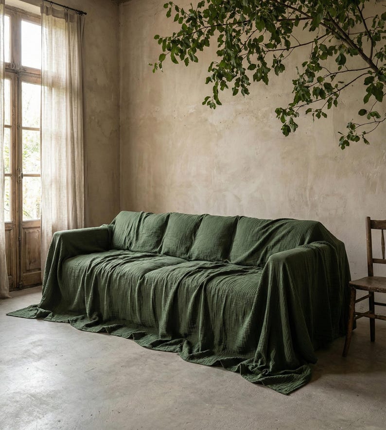 May include: A green sofa draped with a matching green throw. The sofa has several cushions and is positioned in front of a window with sheer curtains. A wooden chair is to the right.