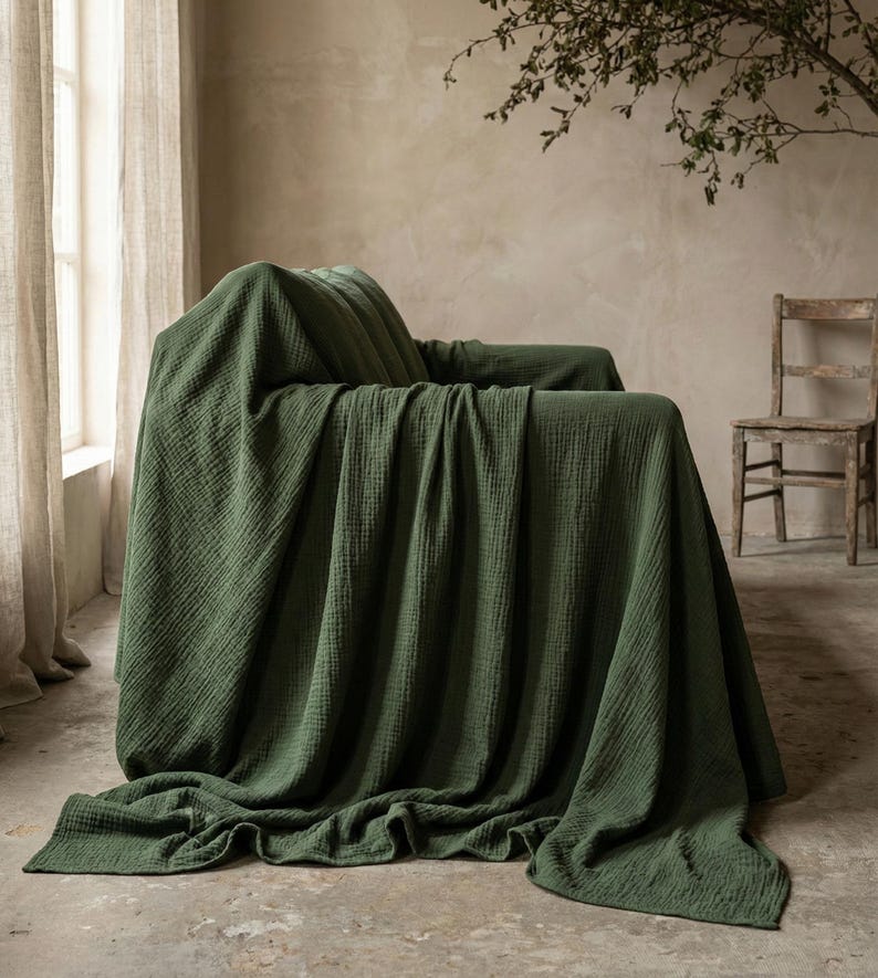 May include: A dark green blanket draped over an armchair. The blanket has a textured appearance and covers the entire chair, with folds cascading to the floor. The room has neutral-toned walls and a wooden chair in the background.