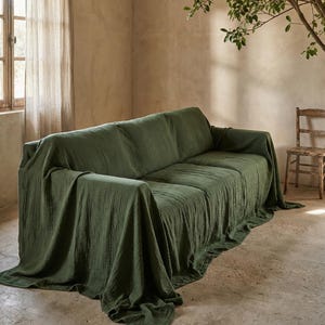 May include: A green sofa draped with a matching fabric throw. The throw cascades over the sofa, forming gentle folds and shadows. The room features a neutral colour scheme, with a window and a wooden chair visible in the background.