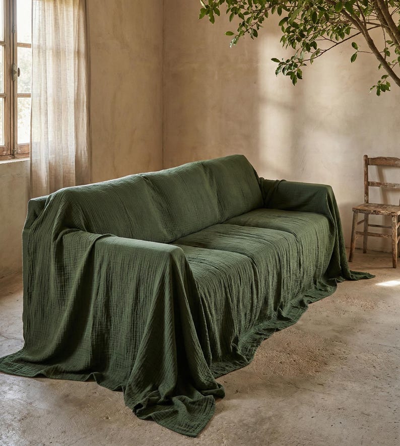 May include: A green sofa draped with a matching fabric throw. The throw cascades over the sofa, forming gentle folds and shadows. The room features a neutral colour scheme, with a window and a wooden chair visible in the background.