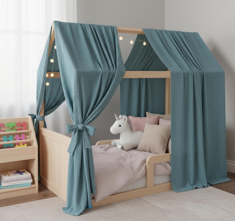 May include: A light wood-framed bed with teal fabric draped over the top, creating a canopy effect. The bed is decorated with pink and beige pillows, a plush unicorn, and a light pink blanket. A wooden bookshelf with colorful blocks is visible.