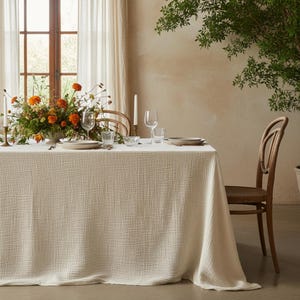 May include: A cream tablecloth drapes a dining table, set for a meal. Plates, glasses, and a floral arrangement with orange and yellow flowers adorn the table. Two wooden chairs are positioned, with a window and sheer curtains in the background.
